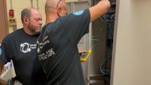 Retail Shrinkage Protection Solutions with Bridge Cable 4 Bridge Cable technicians testing fiber optic and network cabling connections inside a commercial network cabinet.