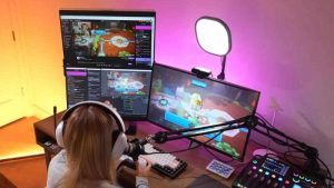 Live streamer wearing headphones using multiple monitors, microphone, lighting, and audio mixer for gaming and online streaming. 