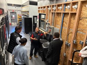Getting Into Low Voltage Careers Through Trade Schools 2 Trade school instructor explaining electrical panels and wiring installations to a group of visitors in an electrical training lab.