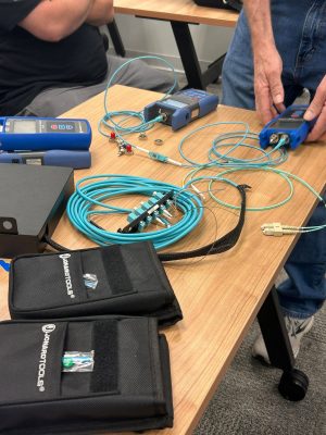 OM5 Fiber for Data Centers and Enterprise Networks 5 Fiber optic technicians using testing equipment and patch cables to measure and certify network fiber connections on a workbench.
