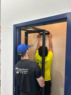 Getting Into Low Voltage Careers Through Trade Schools 3 Two network cabling technicians installing a wall-mounted network rack inside a telecommunications room.