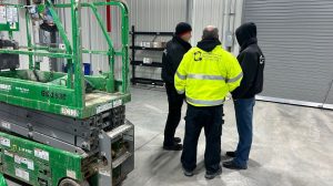 Fiber Optic Contractor: Why They Are Becoming the Standard in Pennsylvania 3 Three technicians, one wearing a Bridgecable Network Cabling high-visibility jacket, standing near a green scissor lift inside a warehouse discussing a project.