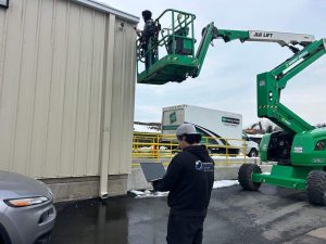 CCTV Contractor: CCTV vs AI Video Analytics in 2026 5 Bridge Cable technician on a boom lift installing an outdoor security camera on a commercial building exterior, while a coworker on the ground monitors progress on a laptop.