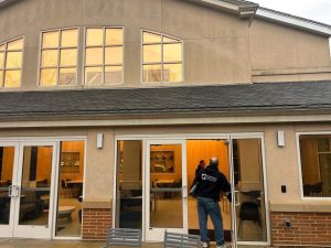 Structured Cabling in Philadelphia: Why Schools Invest? 5 Exterior of a tan stucco building with tall arched windows upstairs, a shingled roof, brick base, and glass entrance doors leading into a lit interior space with tables and chairs; two people stand near the open door.