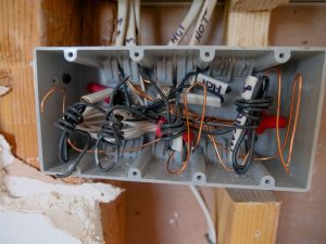 Fiber Optic Contractor Demand Soars This July 1 Open electrical junction box mounted between wood wall studs, showing tangled black, white, and copper wires labeled "HOT" during construction.