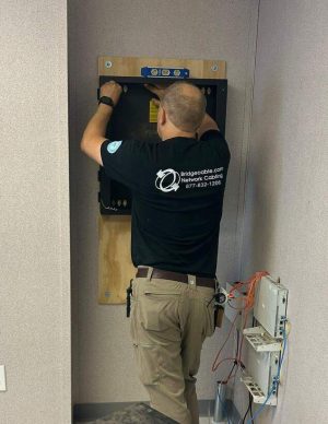 Allentown Structured Cabling Companies: Preparing Networks for Growth 4 Technician in a Bridgecable.com Network Cabling shirt installing a black network cabinet on a wall-mounted plywood backboard, with existing telecom equipment visible nearby.