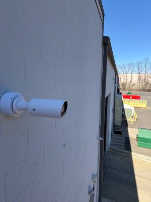 CCTV Contractor: CCTV vs AI Video Analytics in 2026 4 White bullet-style security camera mounted on the exterior cinder block wall of a commercial building, overlooking a parking area with dumpsters and bare trees in the background.