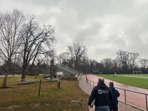 Structured Cabling in Philadelphia: Why Schools Invest? 1 Two Bridge Cable technicians walk alongside an outdoor athletic field and track toward a project site under an overcast sky, carrying equipment for a structured cabling installation.