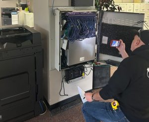 Why Structured Cabling Companies Prevent Service Tickets? 4 A structured cabling companies Technician wearing a black hoodie and tool belt kneeling in front of an open wall-mounted network cabinet, taking a photo of the equipment with a smartphone in a small office setting.