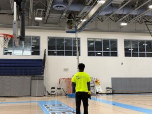 Summer Network Upgrades: WAPs & School Security 3 Bridge Cable technician in a yellow shirt watches from the gym floor as a coworker installs a WiFi access point from a lift near the ceiling.