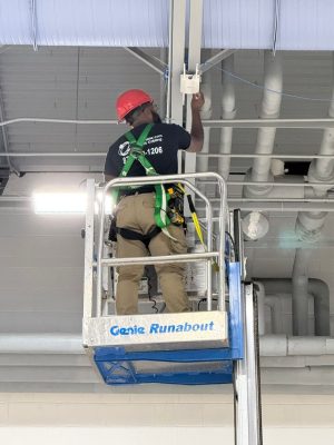 Summer Network Upgrades: WAPs & School Security 4 Bridge Cable technician in a red hard hat and safety harness installs a white dual-antenna WiFi access point from a Genie lift near the ceiling.