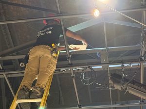 Summer Network Upgrades: WAPs & School Security 2 Bridge Cable technician on a ladder installing a white wireless access point to an overhead beam, with network cabling running along the exposed ceiling.