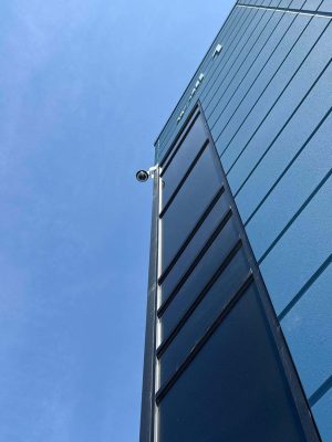 How Weather Extremes Affect Your Security Surveillance 3 Upward view of a bullet camera mounted on the corner of a dark modern building against blue sky.