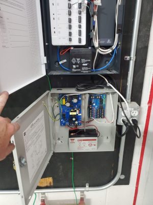 Why Does Access Control During a Power Outage Matter? 5 Open wall-mounted access control cabinet showing a door controller board with trigger lock and REX terminals, a sealed lead-acid backup battery, and a separate Altronix power supply unit with a second 12V battery, connected by conduit to a wall outlet.