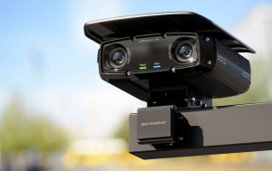 What Role Does Low Voltage Play in Speed Camera Systems? 3 Black AI-powered license plate recognition camera with two stereo-scope lenses, ready and data status lights, and a multi-sensor unit, mounted on a roadside pole against a blue sky.