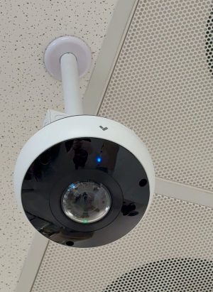 Cloud vs. Local: Which Security Camera Storage Wins? 1 Close-up of a white dome security camera with a black fisheye lens, mounted on a ceiling pole bracket, with blue and green status lights visible, next to a perforated ceiling speaker panel.