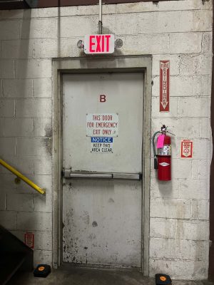 Why Does Access Control During a Power Outage Matter? 2 Gray metal emergency exit door labeled "B" with a horizontal panic bar, lit exit sign above, and a red fire extinguisher mounted on the cinder block wall beside it.