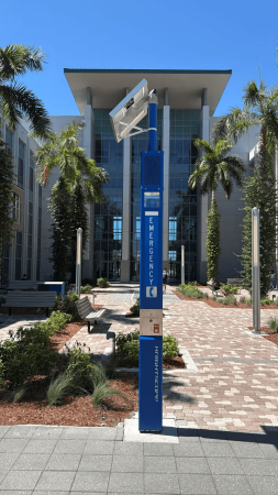 What Are Blue Light Emergency Towers at Hospitals? 3 Blue Knightscope emergency call station with an illuminated "Emergency" panel and red help button, standing on a paved courtyard in front of a modern glass campus building surrounded by palm trees.