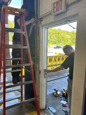 How Do You Choose the Right REX Devices? 3 Two technicians install access control hardware on a commercial exit door: one on a ladder wiring a junction box above the frame near an EXIT sign, the other fitting an electric strike into the door jamb at a loading dock.
