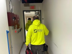 What Is Hybrid Access Control and How Does It Work? 5 Two technicians in yellow Bridgecable.com Network Cabling hoodies walk down an office hallway carrying tool bags, passing a fire alarm panel and an EXIT sign.