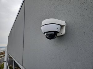 Cloud vs. Local: Which Security Camera Storage Wins? 4 White dome security camera mounted on a textured gray exterior wall of a commercial building, with a wall-mount bracket and clear view of the surrounding parking area and waterfront in the background.