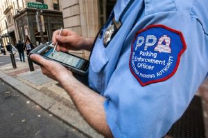 What Role Does Low Voltage Play in Speed Camera Systems? 1 Philadelphia Parking Authority enforcement officer in a blue uniform with a PPA patch entering a citation on a handheld device at a Chestnut Street corner.