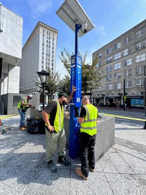 What Are Blue Light Emergency Towers at Hospitals? 4 Three technicians in yellow safety vests install a blue emergency call box topped with a security camera and solar panel on a downtown city sidewalk, with wiring exposed at the base of the pole.