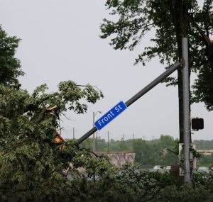 Why Does Access Control During a Power Outage Matter? 1 A blue "Front St" street sign hangs from a bent traffic signal pole, with a large broken tree branch and a yellow vehicle tangled in fallen limbs; a river and low dam are visible in the background.