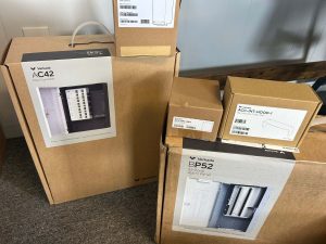 Why Does Access Control During a Power Outage Matter? 4 Cardboard boxes for Verkada access control equipment, including an AC42 four-door controller, a BP52 32-zone alarm panel, a 4AH backup battery, and a rain hood for TD53/TD63 door stations, stacked on a carpeted floor.
